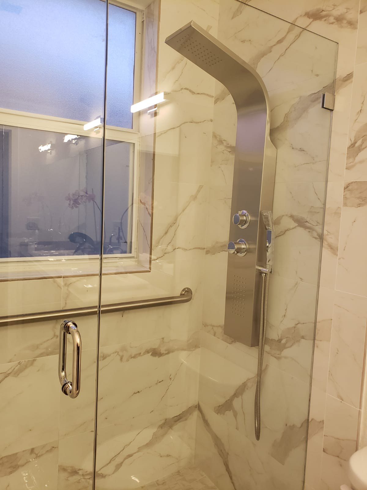 Glass-enclosed shower with beige marble-look tile and window.