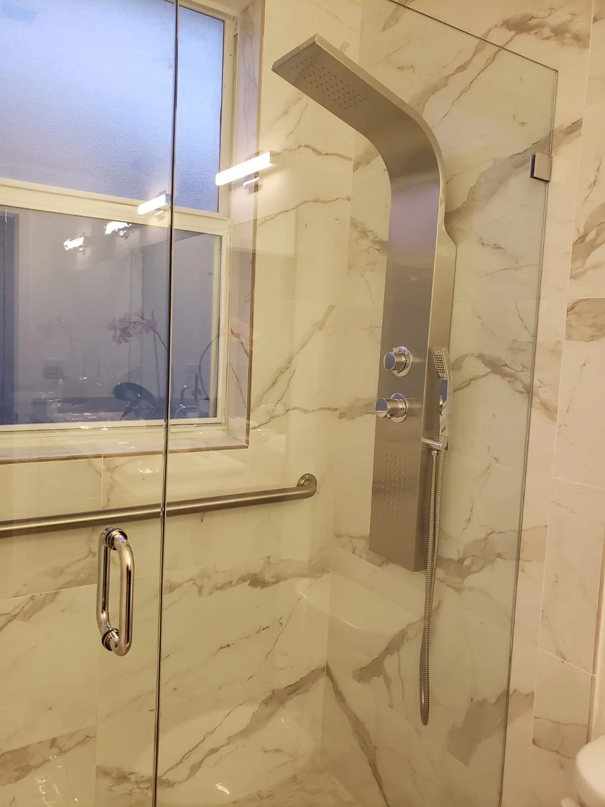 Glass-enclosed shower with beige marble-look tile and window.